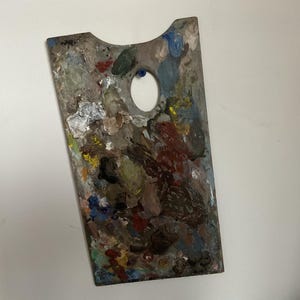 K&ouml;nnte beinhalten: Rechteckige Holzmalpalette mit Daumenloch und gebogenem oberem Rand. Die Palette ist mit einer Vielzahl von getrockneten Farben bedeckt, darunter Rot, Blau, Gr&uuml;n, Gelb und Wei&szlig;. Die Palette wird zum Mischen von Farben verwendet.