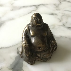 May include: A small, bronze statue of a smiling, seated Buddha. The statue is detailed with folds in the Buddha's robe and a serene expression.