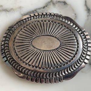 May include: An oval silver belt buckle featuring a central smooth oval plate surrounded by radiating sunburst lines. The outer edge is decorated with a repeating scalloped pattern, creating a textured border around the entire metallic piece.