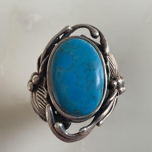 May include: A silver ring with a vibrant, oval turquoise gemstone. The stone is set in a decorative silver setting with leaf and berry details. The ring has a vintage design, suitable for everyday wear or special occasions.