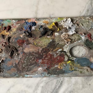 K&ouml;nnte beinhalten: Eine gebrauchte Holzmalpalette, bedeckt mit getrockneter Farbe. Die Palette weist eine Vielzahl von Farben auf, darunter Rot, Blau, Gelb und Gr&uuml;n, mit einem zentralen Daumenloch. Die Palette ist rechteckig mit einer gebogenen Kante.