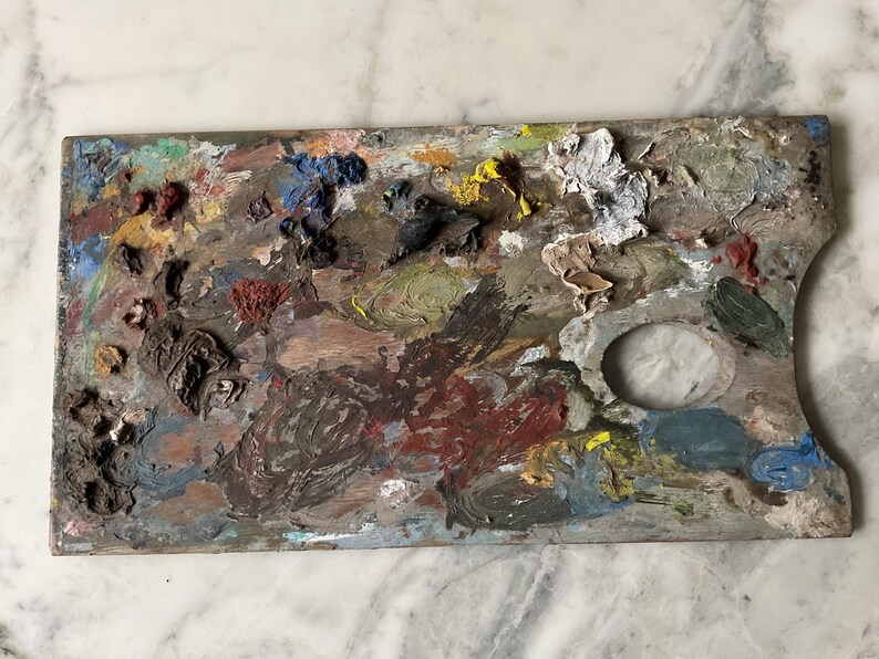 K&ouml;nnte beinhalten: Eine gebrauchte Holzmalpalette, bedeckt mit getrockneter Farbe. Die Palette weist eine Vielzahl von Farben auf, darunter Rot, Blau, Gelb und Gr&uuml;n, mit einem zentralen Daumenloch. Die Palette ist rechteckig mit einer gebogenen Kante.