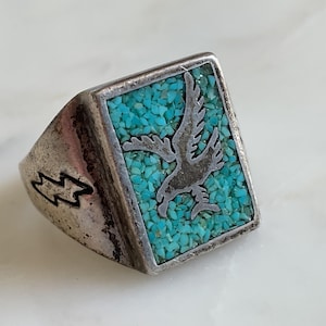 May include: A silver-toned ring featuring a rectangular face inlaid with small, crushed turquoise stones. A metal eagle silhouette is centred within the turquoise inlay, and a lightning bolt symbol is engraved on the side of the ring band.