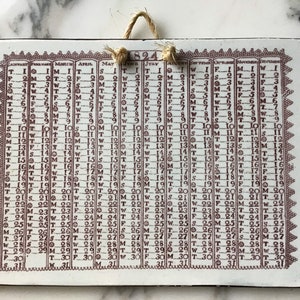 May include: A white ceramic tile calendar with a brown border and brown text. The calendar is for the year 1824 and shows the days of the week and the dates for each month. The tile is hanging from a string.