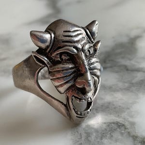 May include: A silver-toned metal ring featuring a sculpted, three-dimensional face of a horned creature with pointed ears, a furrowed brow, and an open mouth. The design includes detailed facial features and a smooth, polished metallic finish.