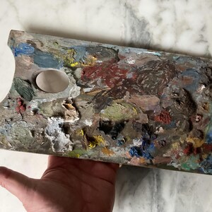 K&ouml;nnte beinhalten: Eine gebrauchte Holzmalpalette, rechteckig mit Daumenloch und ovaler Farbvertiefung. Die Palette ist mit getrockneter Farbe in verschiedenen Farben bedeckt, darunter Rot, Blau, Gelb und Gr&uuml;n. Die Palette wird in einer Hand gehalten.