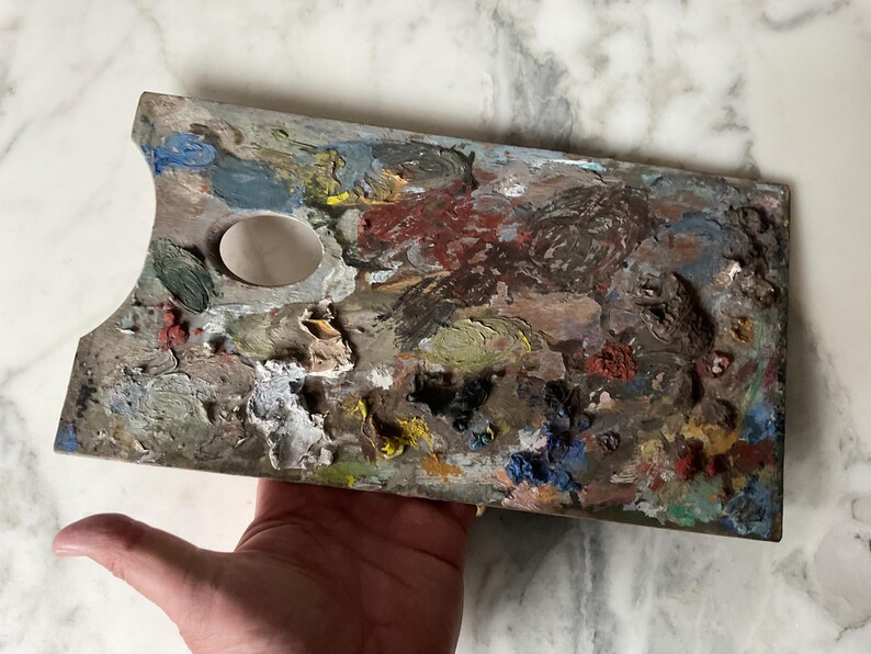 K&ouml;nnte beinhalten: Eine gebrauchte Holzmalpalette, rechteckig mit Daumenloch und ovaler Farbvertiefung. Die Palette ist mit getrockneter Farbe in verschiedenen Farben bedeckt, darunter Rot, Blau, Gelb und Gr&uuml;n. Die Palette wird in einer Hand gehalten.
