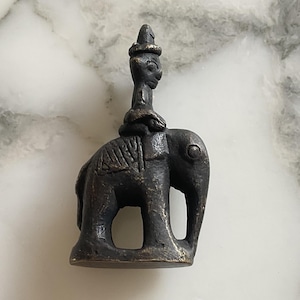 May include: A small, dark gray metal figurine of a person riding an elephant. The figurine is detailed with intricate carvings and is a traditional Asian style.