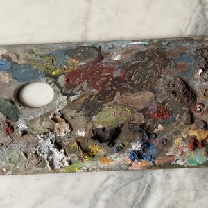 K&ouml;nnte beinhalten: Eine gebrauchte Holzmalpalette, bedeckt mit getrockneter Farbe. Die Palette hat ein Daumenloch und ist stark mit verschiedenen Farben wie Rot, Blau, Gelb und Braun befleckt. Die Palette ist rechteckig mit einer gebogenen Kante.