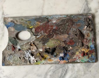 May include: A well-used wooden artist's palette, covered in dried paint. The palette features a thumb hole and is heavily stained with a variety of colors, including red, blue, yellow, and brown. The palette is rectangular with a curved edge.