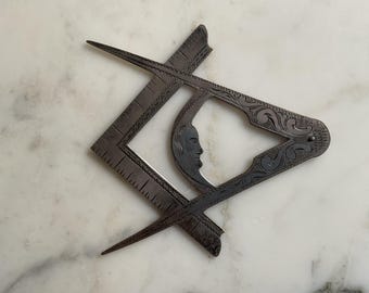 Könnte beinhalten: Ein antikes, dunkles Metall-Freimaurer-Zirkel und -Winkel, mit aufwendigen Gravuren. Der Winkel hat Linealmarkierungen, und der Zirkel zeigt ein Gesichtsprofil. Die Werkzeuge sind in einem symbolischen Layout angeordnet, das Handwerkskunst und Präzision suggeriert.