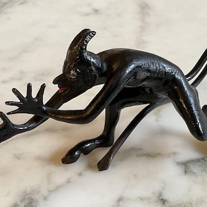 Könnte beinhalten: Eine dunkle, stilisierte Skulptur einer gehörnten Kreatur mit ausgestreckten Händen und einem langen Schwanz. Die Figur hat rote Lippen und scheint sich in einer hockenden Position zu befinden. Die Skulptur steht auf einer Marmoroberfläche.