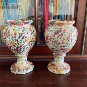 May include: Two white ceramic vases with a colourful splatter paint design. The vases have a round body and a pedestal base.