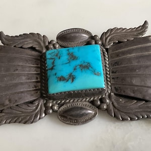 Puede incluir: Broche vintage plateado con una piedra rectangular de turquesa en el centro. El broche presenta un diseño detallado con elementos en forma de hojas y plumas que rodean la piedra. La piedra de turquesa tiene vetas oscuras.