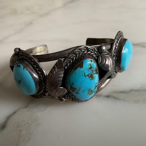 May include: A silver cuff bracelet featuring three oval turquoise stones. The stones are set in ornate, detailed bezels with leaf accents. The bracelet has a vintage or antique aesthetic, with a tarnished finish.