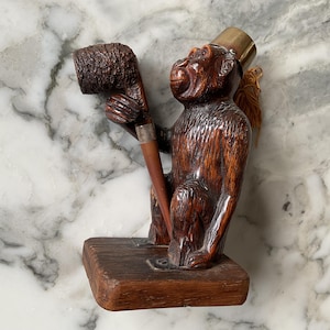 May include: A carved wooden monkey figurine holding a pipe. The monkey is brown with a brass top hat and sits on a wooden base. The pipe is dark brown with a silver band. A decorative item.