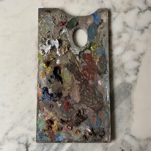 K&ouml;nnte beinhalten: Eine rechteckige Holzmalpalette mit einem Daumenloch. Die Palette ist mit getrockneter Farbe in verschiedenen Farben bedeckt, darunter Wei&szlig;, Gelb, Blau, Gr&uuml;n und Rot. Die Palette liegt auf einer wei&szlig;en Marmoroberfl&auml;che.