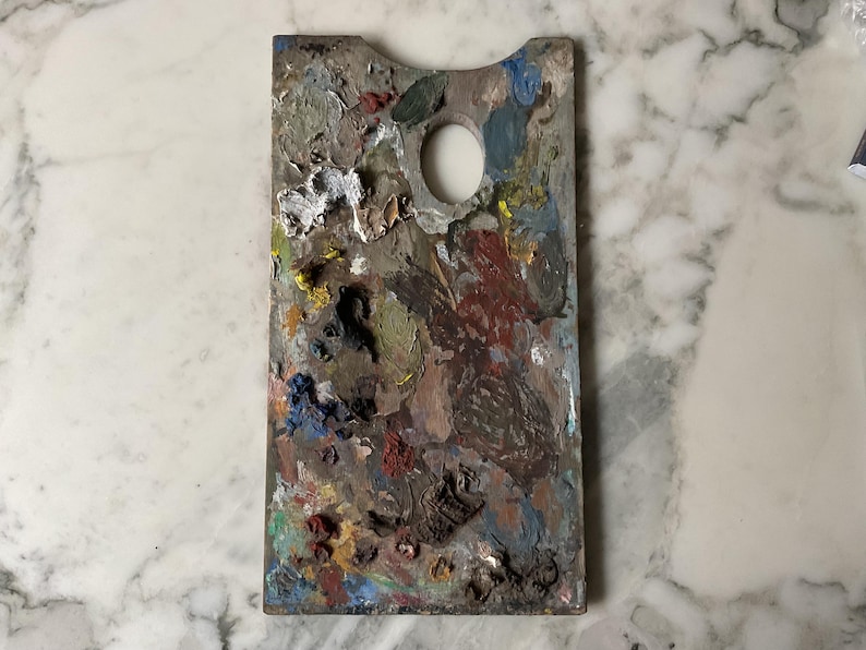 K&ouml;nnte beinhalten: Eine rechteckige Holzmalpalette mit einem Daumenloch. Die Palette ist mit getrockneter Farbe in verschiedenen Farben bedeckt, darunter Wei&szlig;, Gelb, Blau, Gr&uuml;n und Rot. Die Palette liegt auf einer wei&szlig;en Marmoroberfl&auml;che.