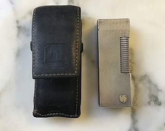 May include: A silver lighter with a textured finish and a black leather case. The case has a stitched edge and a small square logo.