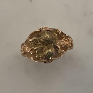 Op de afbeelding: Een gouden ring met een gedetailleerd bladontwerp. De ring is ovaalvormig en heeft een getextureerd oppervlak.