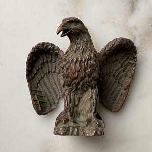 Könnte beinhalten: Eine Bronzeskulptur eines Adlers mit ausgebreiteten Flügeln. Der Adler hat detaillierte Federn und einen leicht geöffneten Schnabel. Die Skulptur hat ein verwittertes Aussehen mit Anklängen von grüner Patina, die auf das Alter hindeuten. Der Adler ist ein dekorativer Gegenstand.