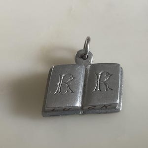 May include: Silver charm in the shape of an open book with the letter "R" engraved on each side. The book is inscribed with the words "LUCK" and "CK".