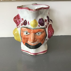 May include: A white ceramic jug with a whimsical design. The jug features a face with a moustache and beard, surrounded by colourful fruit-like shapes.