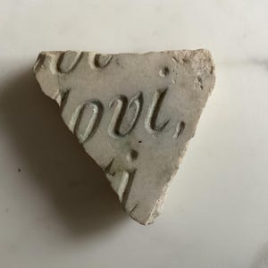 Puede incluir: Fragmento de piedra clara con letras talladas. La palabra "LOVE" es visible, junto con otras letras parciales. La piedra tiene una superficie rugosa y texturizada y tiene forma triangular.