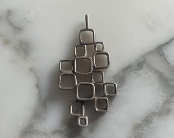 Puede incluir: Colgante plateado con un diseño abstracto de cuadrados interconectados. El colgante tiene una orientación vertical y un pequeño lazo en la parte superior para una cadena. El diseño es geométrico y moderno, adecuado para el uso diario.