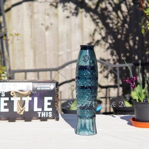 May include: A blue glass vase shaped like a fish with a textured surface. The vase is sitting on a white table with a box of Guinness beer in the background.
