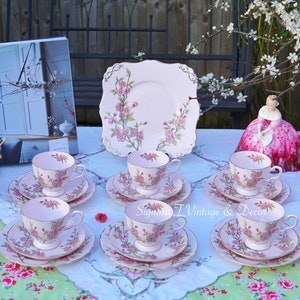 May include: A set of six pink teacups and saucers with a delicate floral design. The cups and saucers have a gold trim and are arranged on a floral tablecloth. A square pink cake plate with a matching floral design is in the centre of the table.