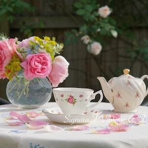 May include: A white teapot and teacup with pink rose patterns are on a white tablecloth with pink rose petals scattered around them. A bouquet of pink roses in a clear vase is on the table.