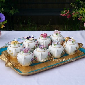 May include: A set of eight miniature porcelain teapots with floral designs and 3D flower accents. The teapots are arranged on a teal and gold tray. Signora T Vintage & Decor.