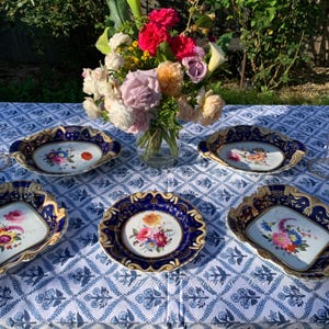 May include: A collection of antique porcelain serving dishes with a dark blue and gold trim, each featuring a hand-painted floral design. A vase of colorful flowers sits in the background on a blue and white patterned tablecloth.