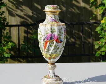 May include: A white porcelain vase with a gold trim and a lid. The vase is decorated with a colorful bird and floral pattern. The vase is on a white surface.