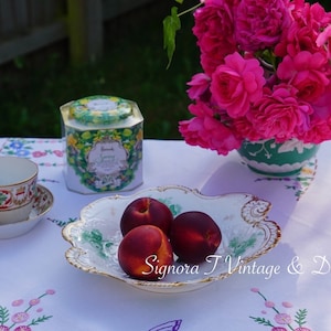 May include: A white tablecloth with embroidered flowers, a white teacup and saucer with floral designs, a green and white tea tin with the text "Brooke Bond Sunny Breakfast", a white porcelain bowl with green and gold accents, and three red peaches.