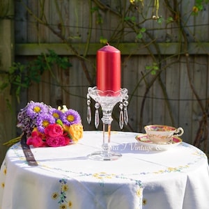 Puede incluir: Un candelero de cristal con una vela roja sobre un mantel blanco con un ramo de flores moradas, rosas y amarillas. El candelero tiene detalles de cristal en forma de lágrima.