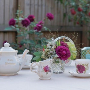 May include: White tea set with a teapot, creamer, and two teacups and saucers. The teapot has gold trim and the text "Ruby Wedding" on the side. The teacups and saucers have floral designs with pink roses.