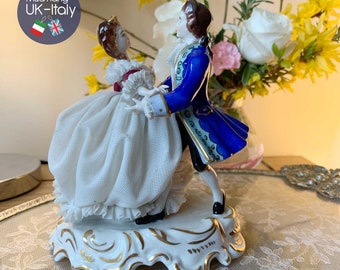 May include: A porcelain figurine of a couple dancing. The woman is wearing a white dress with lace trim and the man is wearing a blue coat with gold accents. The figurine is on a white base with gold trim.