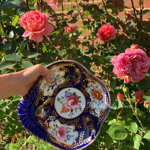 May include: A blue and gold porcelain dish with floral designs. The dish is decorated with gold trim and has a scalloped edge. The dish is being held in a hand in front of a background of pink roses and green leaves.