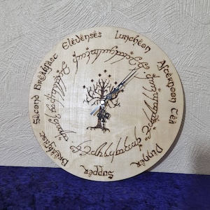 Puede incluir: Un reloj de madera con un diseño de árbol de la vida y la inscripción "El Anillo Único" en escritura élfica. La esfera del reloj está dividida en secciones para cada comida del día, incluyendo "Desayuno", "Segundo Desayuno", "Once", "Almuerzo", "Té de la tarde", "Cena" y "Cena".