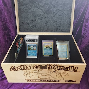 May include: A wooden box with a black velvet interior and a Pokemon card game theme. The box has a wood-burned image of a Pokemon character and the text "Gotta catch 'em all!" The box is open and contains three Pokemon cards.