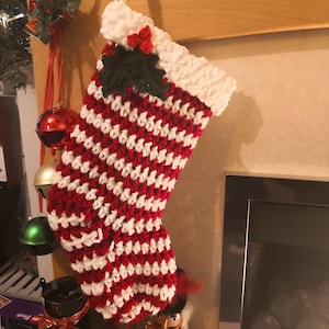 Puede incluir: Una media de Navidad de crochet roja y blanca con una rama de acebo verde y bayas rojas en la parte superior. La media está colgada en una repisa de chimenea.