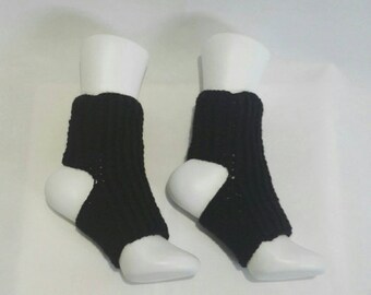May include: Two black knitted leg warmers on white mannequins. The leg warmers are ribbed and have a heel cutout. The background is a plain white surface.