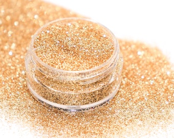 May include: A clear, small plastic jar filled with gold glitter, sitting on a bed of loose gold glitter. The glitter sparkles and reflects light, creating a shimmering effect. The background is white, highlighting the glitter's color and texture.