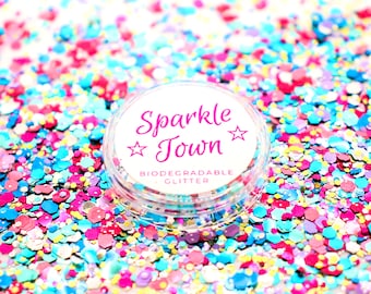 May include: A small clear jar of biodegradable glitter with a white label that says "Sparkle Town" in pink lettering with stars on either side of the word "Town". The jar is surrounded by colorful glitter.