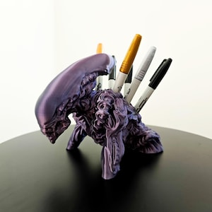 Puede incluir: Un portalápices morado con forma de criatura alienígena, con bolígrafos insertados en la parte posterior. El portalápices es una escultura detallada con una superficie texturizada. Los bolígrafos son dorados, plateados, negros y naranjas.
