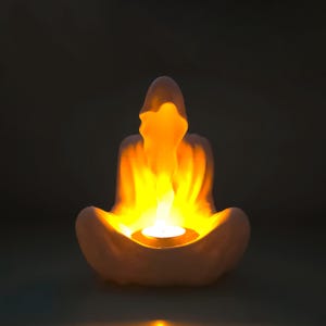 May include: A white ceramic candle holder shaped like a person sitting in a meditative pose. The candle holder has a small opening in the center for a tea light candle. The candle is lit and glowing.