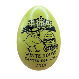 Puede incluir: Un objeto de colección en forma de huevo verde claro con una ilustración negra de la Casa Blanca, un pollito empujando un huevo decorado en un carrito y el texto "WHITE HOUSE EASTER EGG ROLL 2000".