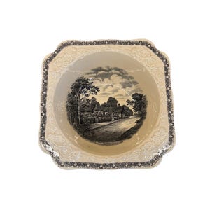 May include: Cream-coloured, square ceramic bowl with a black and white landscape scene. The rim has a decorative, scalloped edge with a black floral pattern. The bowl's interior features a detailed illustration of a village scene.